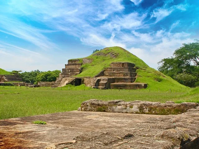 Antiguas ruinas mayas con montículos cubiertos de hierba y estructuras de piedra bajo un cielo azul